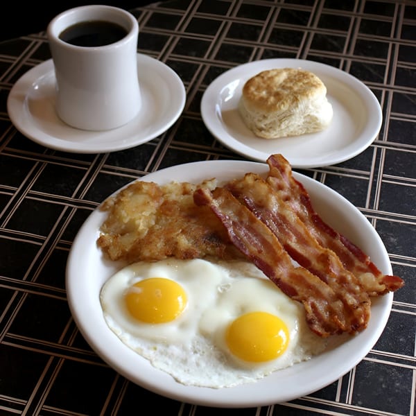 A classic breakfast of fried eggs, bacon, hash browns, a biscuit, and coffee on a checkered table.
